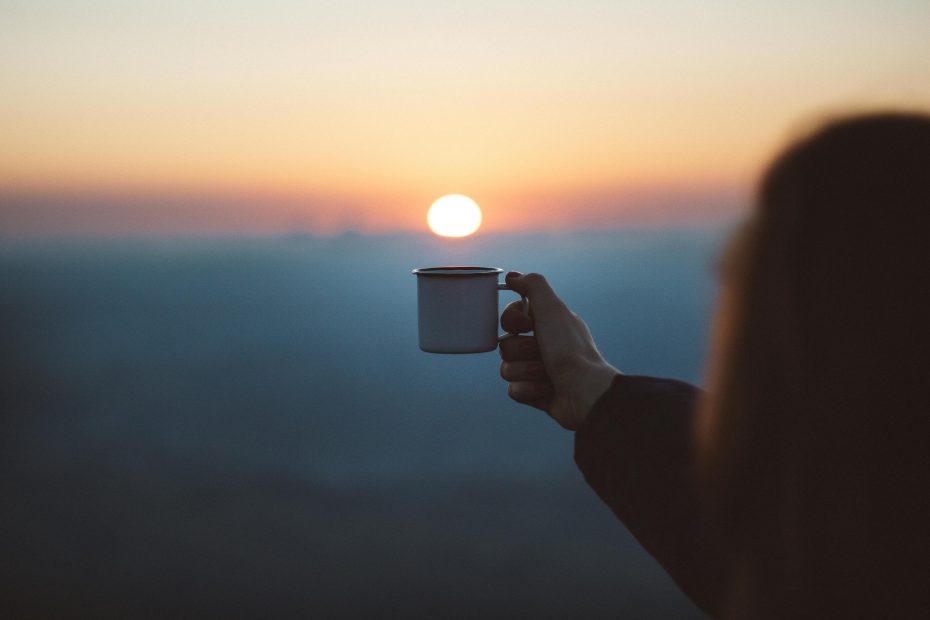 A cup of coffee is held in the forefront while the sun rises in the background.
